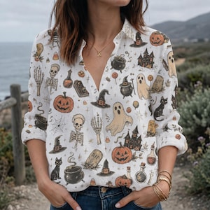 May include: A white long-sleeve button-down shirt featuring a repeating Halloween-themed pattern. The print includes orange pumpkins, ghosts, black cats, witch hats, cauldrons, skeletons, tombstones, and haunted houses in a detailed illustration style.