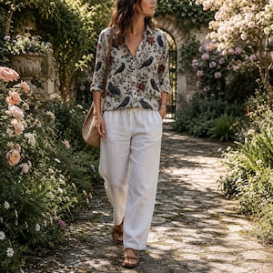 May include: A long-sleeved button-down shirt featuring a pattern of black crows and red flowers on a light gray background, paired with loose-fitting white trousers and a woven straw shoulder bag. The shirt has a collar and a front button placket.
