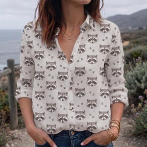May include: A long-sleeved button-down shirt in a light beige color featuring an all-over repeating pattern of grey and black raccoon faces interspersed with small black paw prints. The shirt has a classic collar and a front button closure.