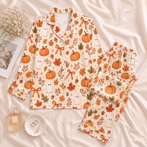 Könnte beinhalten: Ein langärmeliges Schlafanzug-Set mit Knopfleiste und passender Hose in Cremefarben, das mit einem durchgehenden Muster aus orangefarbenen Kürbissen, weißen Geistern, braunen Eicheln, Herbstblättern und orangefarbenen Schleifen bedruckt ist.