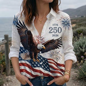 Puede incluir: Una camisa blanca de manga larga con botones que presenta un gráfico de un águila calva en vuelo sobre una bandera estadounidense. El diseño incluye patrones de fuegos artificiales rojos, blancos y azules y el texto 1776, 250 y 2026 a la derecha.