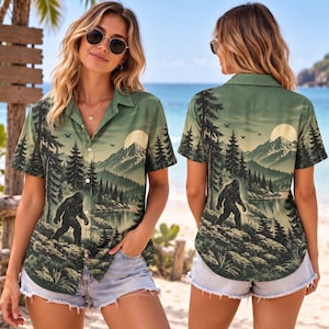 May include: A short-sleeved button-down shirt featuring a green-toned landscape print of a forest, mountains, a lake, and a large creature resembling Bigfoot. The design includes a full moon in the sky and is displayed from both front and back views.