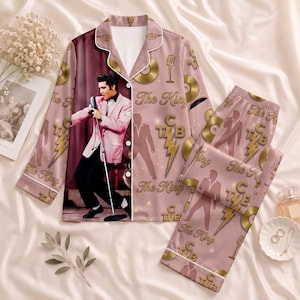 May include: A dusty pink pajama set featuring a long-sleeve button-down shirt and matching pants. The fabric displays a repeating pattern of gold lightning bolts, microphones, vinyl records, and the text "The King" and "TCB" alongside a large image of Elvis.