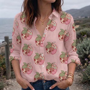 May include: A light pink long-sleeve button-down shirt featuring a repeating pattern of apples. Each apple graphic is composed of a patchwork design with various green, red, and white checkered and floral prints. The shirt has a classic collar.