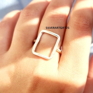 Puede incluir: Un anillo de plata con un diseño rectangular abierto. El anillo se lleva en un dedo, y el diseño es una forma simple y moderna. El texto "SILVERARTGIFT25" es visible en la imagen.