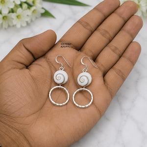 May include: A pair of dangle earrings featuring a white spiral shell bead attached to a silver-toned hammered metal hoop. The earrings hang from silver-toned fishhook wires. The shell beads display a natural spiral pattern in shades of white and tan.