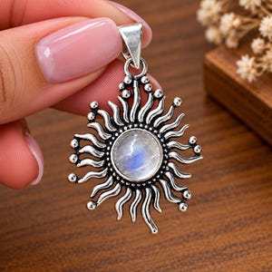 May include: A silver-toned metal pendant featuring a central round moonstone cabochon surrounded by radiating, wavy sunburst rays. The metalwork has a darkened, oxidized finish in the recessed areas, highlighting the textured, curved rays.
