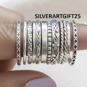 May include: A collection of silver rings with various designs, including braided, patterned, and plain bands. The rings are displayed on a finger, showcasing their different textures and styles. The text "SILVERARTGIFT25" is visible.