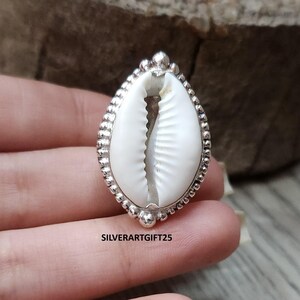 Peut inclure: Bague en argent ornée d'un coquillage porcelaine blanc. La bague présente un bord perlé détaillé et une finition polie. Le texte "SILVERARTGIFT25" est visible.