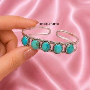 May include: A silver cuff bracelet featuring five oval-shaped turquoise stones set in rope-textured bezels. The band has an open design with etched detailing along the metal surface. The text SILVERARTGIFT25 is visible above the bracelet.
