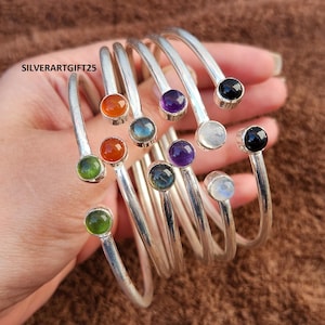 以下が含まれることがあります： さまざまな宝石のアクセントが付いたシルバーブレスレット。緑、オレンジ、青、紫、白、黒の石が含まれています。ブレスレットは手の中にあり、さまざまな色とデザインが示されています。「SILVERARTGIFT25」の文字が見えます。