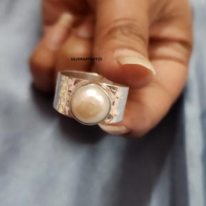 Puede incluir: Un anillo de plata con una perla grande, redonda y blanquecina en el centro. La banda tiene una textura martillada. El anillo se sostiene en una mano sobre un fondo azul. El texto "SILVERARTGIFT25" es visible.