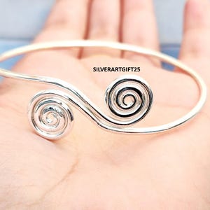 May include: A silver bracelet with a spiral design. The bracelet has two spiral features, one at each end of the open cuff. The text "SILVERARTGIFT25" is visible on the bracelet.