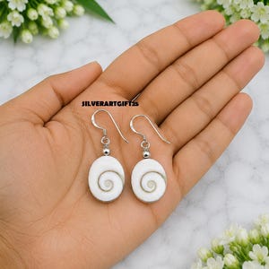 May include: A pair of dangle earrings featuring oval-shaped white Shiva eye shells with a natural spiral pattern. Each shell is attached to a silver-toned metal hook by a small silver-toned bead. The text SILVERARTGIFT25 is visible in the center.