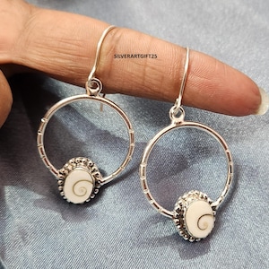 Op de afbeelding: Een paar zilveren oorringen met een wit, ovaalvormig middenstuk met een spiraalvormig ontwerp. De oorbellen hebben een haaksluiting. De tekst "SILVERARTGIFT25" is zichtbaar. Gepresenteerd tegen een blauwe achtergrond.