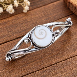 Puede incluir: Un brazalete de metal plateado con una concha de ojo de Shiva blanca circular en el centro que presenta un patrón de espiral natural. La banda está hecha con alambre de metal pulido que se curva en bucles abiertos y pequeños acentos esféricos.