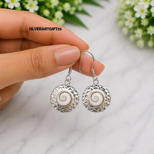 May include: A pair of silver dangle earrings featuring a circular, hammered metal disc with a central white and brown spiral shell inlay. The earrings hang from simple silver hooks and include the text SILVERARTGIFT25 printed on the surface.