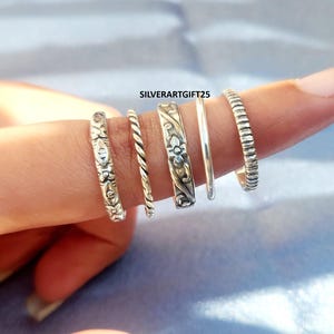 May include: Five silver rings of varying designs are displayed on a finger. The rings include floral patterns, a twisted rope design, and a ribbed band. The text "SILVERARTGIFT25" is visible. These rings are suitable for everyday wear.