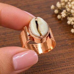 Op de afbeelding: Een ring met een gepolijste koperkleurige metalen band met een breed, plat profiel. Het middelpunt is een natuurlijke, gebroken witte kauri-schelp die stevig is gemonteerd in een bijpassende koperkleurige metalen zetting voor een contrast.