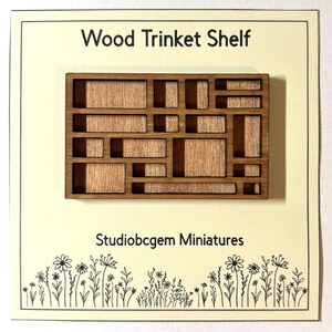 May include: A miniature wooden trinket shelf with a geometric design. The shelf is made of light brown wood and features various rectangular compartments. The text "Wood Trinket Shelf" and "Studiobcgem Miniatures" are visible.