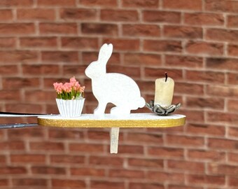 May include: A white bunny silhouette figurine, a small potted pink flower, and a white candle on a gold shelf.