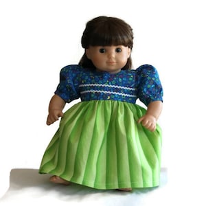May include: A doll dressed in a blue floral top with short, puffed sleeves and a green and white striped skirt. The doll has brown hair and is set against a white backdrop. The dress features white trim.