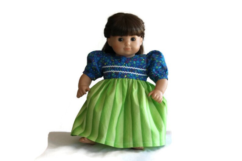 May include: A doll dressed in a blue floral top with short, puffed sleeves and a green and white striped skirt. The doll has brown hair and is set against a white backdrop. The dress features white trim.