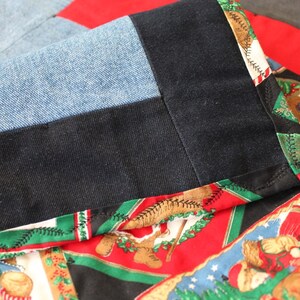 May include: Close-up of a quilt made from different fabrics, including denim, black fabric, and a red fabric with a Christmas-themed print. The quilt is sewn together with a green and black trim.