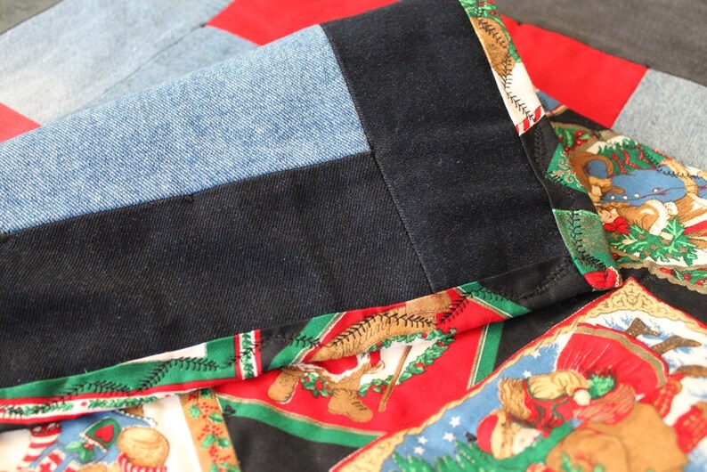 May include: Close-up of a quilt made from different fabrics, including denim, black fabric, and a red fabric with a Christmas-themed print. The quilt is sewn together with a green and black trim.