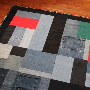 May include: A patchwork quilt made from denim fabric in various shades of blue, black, and red. The quilt is made up of squares and rectangles of different sizes, and the seams are visible. The quilt is edged with a black fabric.
