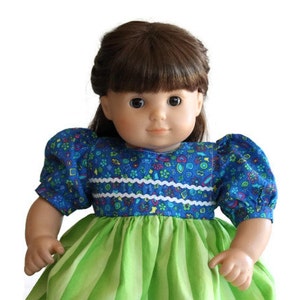 May include: A doll with brown hair and eyes wearing a blue floral print top with white trim and green and white striped skirt. The doll has a fair complexion and is set against a white background.