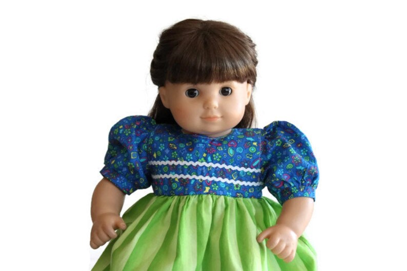 May include: A doll with brown hair and eyes wearing a blue floral print top with white trim and green and white striped skirt. The doll has a fair complexion and is set against a white background.