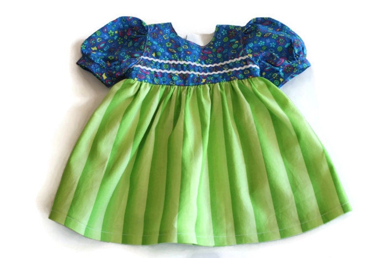 May include: A baby dress featuring a blue floral patterned bodice and short puffed sleeves. The bodice is trimmed with white. The skirt is bright green with vertical stripes. The dress is made of soft fabric.