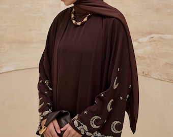 May include: A dark brown abaya with gold embroidery featuring crescent moons and stars on the sleeves. The abaya is paired with a matching head covering and a beaded necklace. A black clutch is held in the hands.