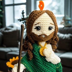 Könnte beinhalten: Handgefertigte Häkelpuppe, die eine religiöse Figur darstellt, mit braunem Bart und Haaren, einem grünen Schal und einem weißen Gewand. Die Puppe hält einen Holzstab und hat einen orangefarbenen Heiligenschein. Die Puppe ist etwa 25 cm groß.