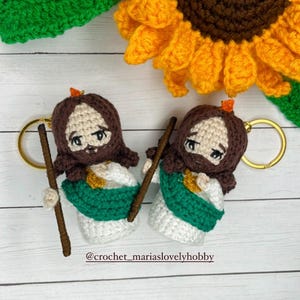 May include: Two crochet keychains depicting a figure with brown hair and a beard, wearing a white robe with a green sash. Each keychain has a gold ring and a wooden staff. The background includes a crochet sunflower.