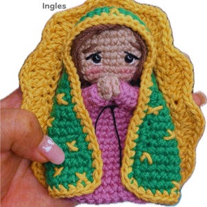 May include: A handmade crochet doll of the Virgin Mary, featuring a pink dress, green and yellow shawl, and a yellow halo. The doll has a sweet expression and is held in a hand. Text on the image reads "PDF Español/Ingles" and "Maria's lovelyhobby."