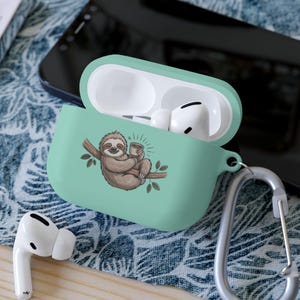 May include: A mint green AirPod case with a cartoon sloth design. The sloth is brown and holding a cup of coffee while sitting on a tree branch. The case is open, revealing white AirPods inside. A silver carabiner clip is attached.