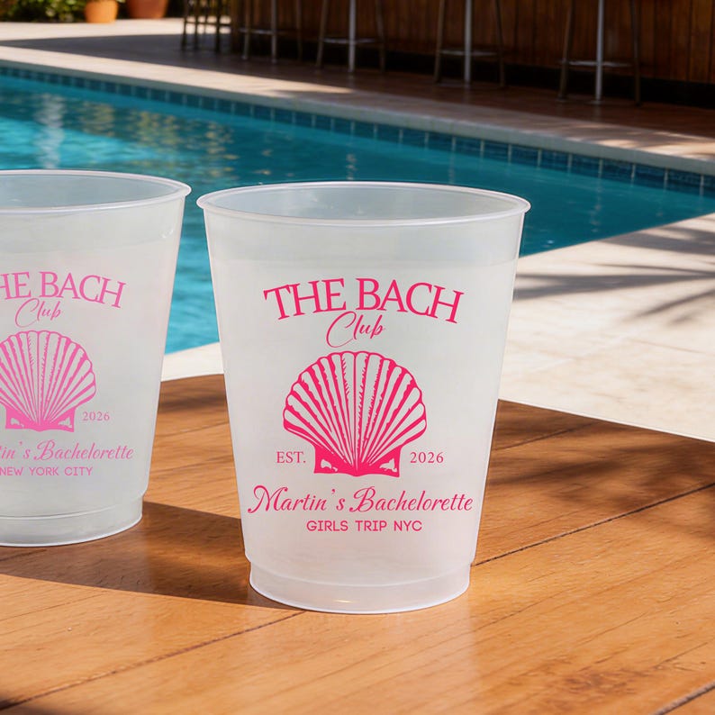 Puede incluir: Dos vasos de pl&aacute;stico transl&uacute;cido esmerilado con texto en rosa intenso y un gr&aacute;fico de concha marina. La impresi&oacute;n dice THE BACH Club, EST. 2026, Martin's Bachelorette y GIRLS TRIP NYC. Los vasos est&aacute;n colocados sobre una superficie de madera.