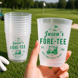 May include: A stack of clear plastic cups with green printed text and graphics. The design features a golf cart, a flag, and the text "Jason's FORE-TEE EST. 1986 GOOD TIMES GREAT SWINGS". One cup is held in the foreground, showing the design clearly.