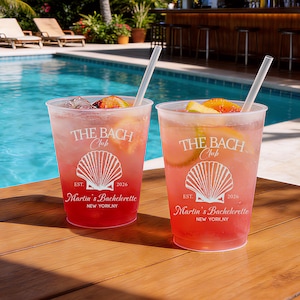 May include: Two clear plastic cups filled with a pink cocktail, ice, and citrus slices, each featuring a white straw and a printed design of a seashell with the text THE BACH Club, EST. 2026, Martin's Bachelorette, and NEW YORK, NY.