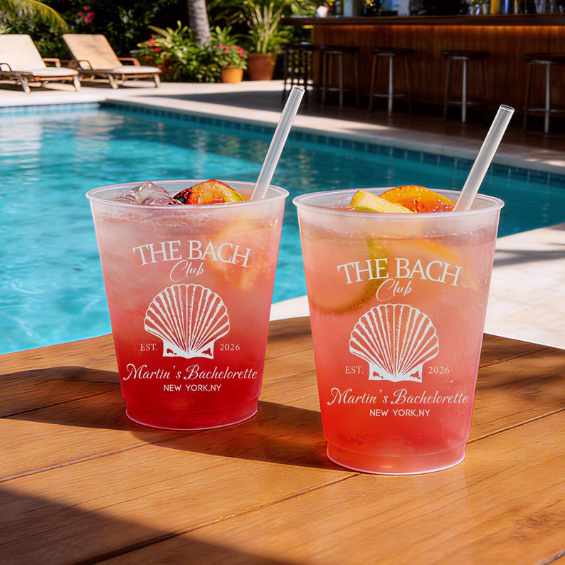 May include: Two clear plastic cups filled with a pink cocktail, ice, and citrus slices, each featuring a white straw and a printed design of a seashell with the text THE BACH Club, EST. 2026, Martin's Bachelorette, and NEW YORK, NY.