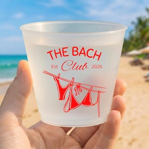 May include: A translucent frosted plastic cup featuring red printed text that reads THE BACH CLUB Est 2026 above a graphic of two bikini pieces hanging from a clothesline with clothespins, set against a blurred beach background.