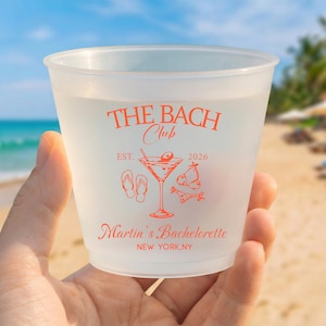 May include: A frosted plastic cup featuring orange text and graphics. The design includes the words THE BACH CLUB, EST. 2026, Martin's Bachelorette, and NEW YORK, NY, alongside illustrations of a martini glass, flip-flops, and swimwear.
