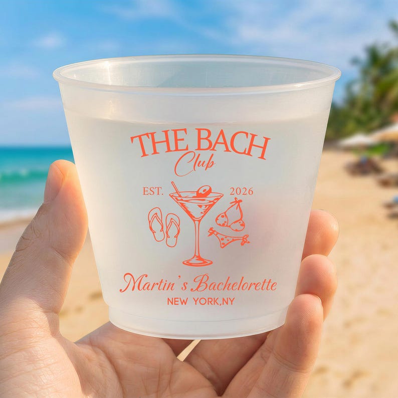 May include: A frosted plastic cup featuring orange text and graphics. The design includes the words THE BACH CLUB, EST. 2026, Martin's Bachelorette, and NEW YORK, NY, alongside illustrations of a martini glass, flip-flops, and swimwear.