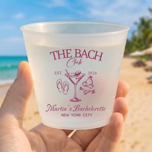 Puede incluir: Un vaso de pl&aacute;stico esmerilado con texto y gr&aacute;ficos de color granate. El dise&ntilde;o incluye las palabras THE BACH Club, EST. 2026, Martin's Bachelorette y NEW YORK CITY, junto con ilustraciones de una copa de martini, chanclas y un bikini.