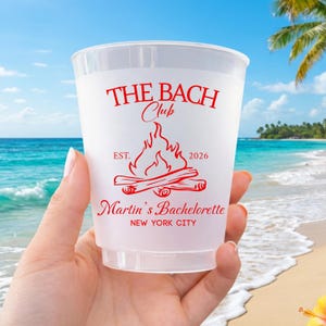 May include: A frosted plastic cup featuring red text and a campfire graphic. The text reads THE BACH Club, EST. 2026, Martin's Bachelorette, and NEW YORK CITY. The design is printed in a bold, stylized font with a central bonfire illustration.
