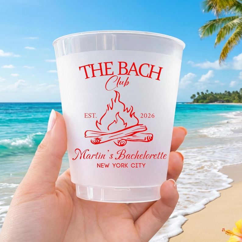 May include: A frosted plastic cup featuring red text and a campfire graphic. The text reads THE BACH Club, EST. 2026, Martin's Bachelorette, and NEW YORK CITY. The design is printed in a bold, stylized font with a central bonfire illustration.