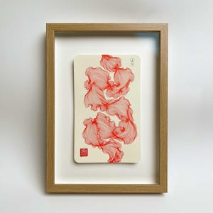 Könnte beinhalten: Ein gerahmtes Kunstwerk. Eine cremefarbene rechteckige Karte zeigt ein rotes, fließendes, abstraktes Design. Das Kunstwerk ist in einem hellbraunen Holzrahmen vor einem weißen Hintergrund eingefasst. Das Design enthält kleine Symbole und einen roten Stempel.
