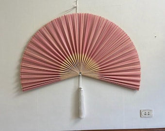 May include: A large, semi-circular wall fan featuring pleated pink paper with a light beige center radiating outward. The fan is suspended by a thin white string and includes a long, white decorative tassel hanging from the central pivot point.